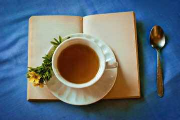 Warm cup of tea placed on an open book with sprig of yellow flowers, set on vibrant blue tablecloth