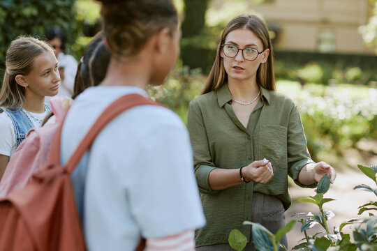 Young female teacher wearing eyeglasses speaking about plants during outdoor class in botanical garden