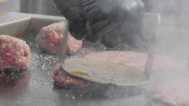 A chef in black gloves prepares a trendy smash burger on a flat griddle, close-up. Juicy meat and melted cheese look appetizing. Street food at a city festival.