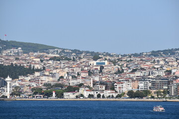 View of the Bosphorus with a densely packed hillside covered in residential buildings, a large Turkish flag on the hill, and boats on the water under a clear blue sky
