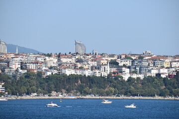 View of the Bosphorus with boats on the water, a residential hillside, and modern skyscrapers in the background under a clear blue sky
