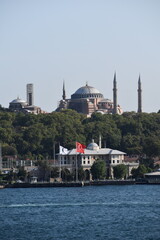 Obraz premium View of Hagia Sophia with Turkish and Yeşilay flags in the foreground along the Bosphorus in Istanbul, framed by lush greenery and the waterfront on a sunny day 