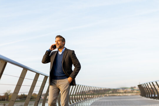 A businessman walking on a modern bridge in the city while talking on his phone during a clear afternoon - Powered by Adobe