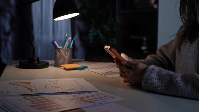 Adult business woman sitting at desk working late at night in dark office, watching scrolling social media on smartphone, procrastinating work.