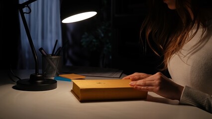 Obraz premium Adult business woman sitting at desk working late at night in dark office, looking at book, holding hands on the cover, late night self education.