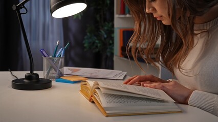 Adult business woman sitting at desk working late at night in office, reading a book, holding hand...