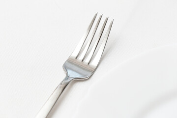 Single silver fork on a white table cloth next to a plate. Closeup of elegant dishware in a luxury dining setting. Minimalist table arrangement with clean, shiny cutlery background.