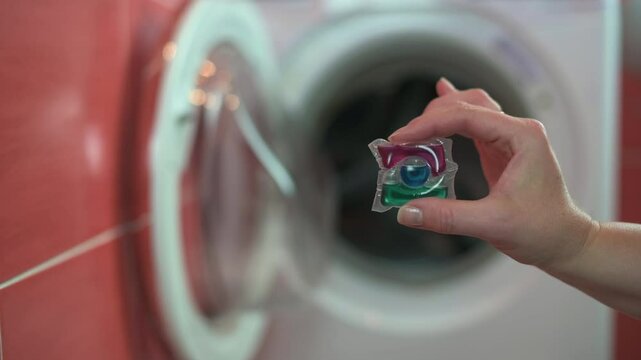 hand holding a colorful laundry detergent pod in front of an open washing machine, ready for use. Focus on the pod with a blurred appliance background.