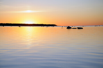 A serene sunset over calm waters with soft golden hues reflecting on the surface. Silhouetted rocks and a distant treeline add depth to this tranquil landscape.