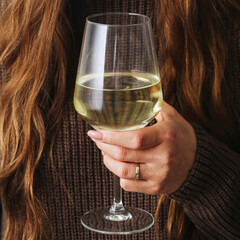 Woman holding a wine glass and sipping white wine. Cozy winter vibes with woolen sweater. Female enjoying a drink during the cold season. Relaxed lifestyle and seasonal alcohol drinking.