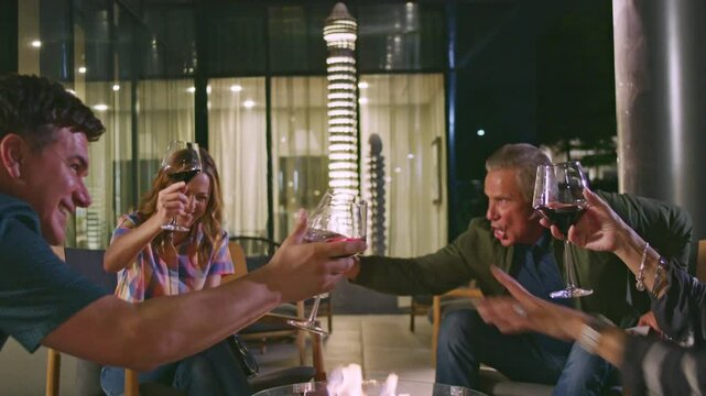 A group of friends cheers with wine glasses around a firepit in a modern setting