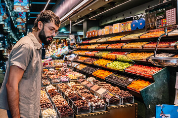 Bearded man choosing sweets at market stall with variety of candies and dried fruits