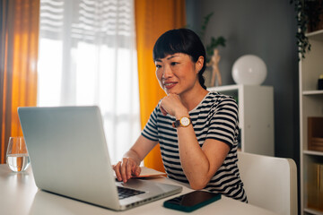 Asian woman working diligently on laptop at a well-lit home office.