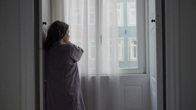 Unhappy or sad woman closing a window curtain overlooking the city street. Feeling melancholic or wanting time alone she pulls down white fabrics to block the sunlight from entering room or apartment