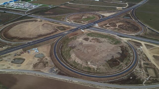 Top-down drone view of a newly built highway cloverleaf interchange with looping ramps, overpasses and active motorway traffic. Large-scale transportation infrastructure and civil engineering project 