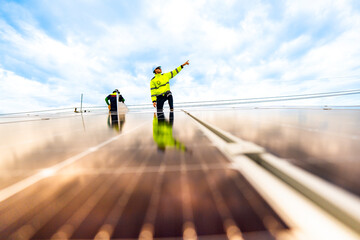 A technician in safety gear inspects solar panels on a metal rooftop, holding a tool. The scene...
