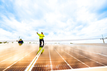 A technician in safety gear inspects solar panels on a metal rooftop, holding a tool. The scene...