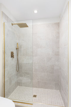 A walk-in shower with square brown tiles, glass wall, and a gold shower head, shower hose, and drain.