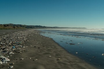 Obraz premium Pollution Crisis on the Beach: Plastic Debris Accumulates Along the Shoreline in a Scenic Coastal Landscape Under Clear Blue Sky