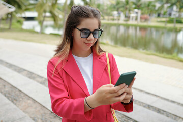 Fototapeta premium Woman wearing sunglasses and pink blazer using smartphone while standing on sidewalk by a pond, smiling and seeming content, with green trees in background