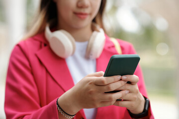 Asian woman wearing headphones and holding a smartphone, standing outdoors in a casual setting. She is engaging deeply with her phone