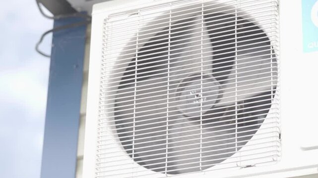 A close-up view of an air conditioning fan is presented. The outdoor air conditioning unit is mounted on the wall of a house. The fan features a white body with a blue and white logo. The fan appears