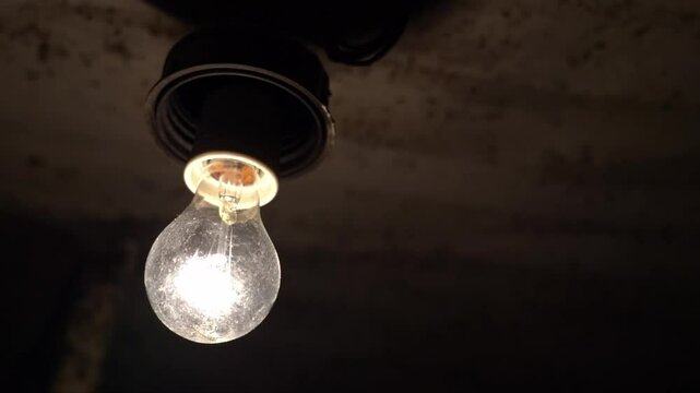 A close-up on a black background shows an incandescent lamp that turns on and off, creating a flickering effect. The filament, emitting a warm light, seems to dance in the wind. A light bulb in a dark