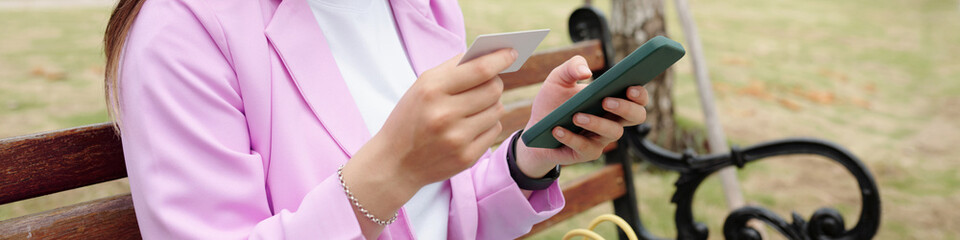 Person holding smartphone and credit card while sitting on park bench, wearing pink blazer with white shirt. Background includes park greenery and bench details