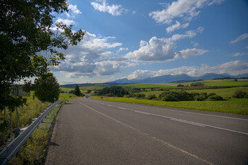 Scenic countryside road surrounded by lush green fields and distant mountains under a sunny sky.