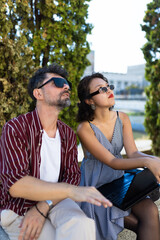 Obraz premium Young man and woman wearing sunglasses sitting outdoors with laptop and looking up on a sunny day.