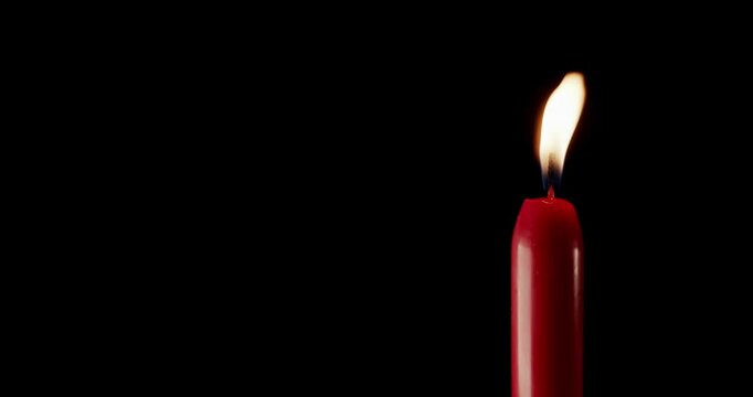 A single red candle burning. Isolated candle burning with dark background. Red paraffin candle with yellow shades burns on a black background. Background or illustration of remembrance or celebration.