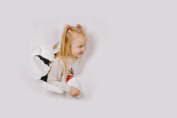 A smiling young girl with blonde hair peeks through a torn white paper wall, wearing a festive sweater. Bright white background and copy space.