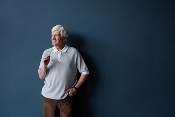 Minimal portrait shot of white haired senior man wearing casual outfit looking away standing on blue wall in studio with natural light and holding glasses