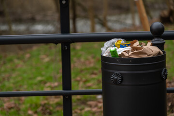 Close-up side view of black metal garbage bin full of mixed litter in autumn public park in cloudy...
