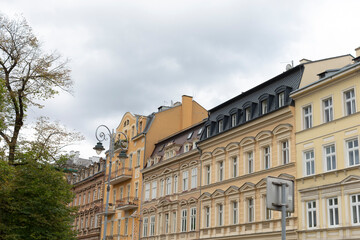 In the scene, there is a row of diverse buildings that are set against a backdrop of a cloudy sky, with lovely trees filling the foreground