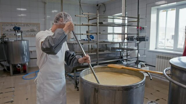 Medium full shot of young Caucasian male cheese factory technician standing by large metal heating vat and stirring raw fermenting milk mass with large whisk during coagulation