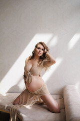 Fototapeta premium A beautiful pregnant girl dressed in Afro style, with loose hair and pigtails sits in a beige beach suit on a sofa by the window. Wooden bracelets on the arm, beautiful makeup. A woman waiting child