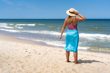 Obraz premium Summer vacation. Tanned mature woman in swimsuit, pareo and sun hat standing by shore on sandy beach. Back view 