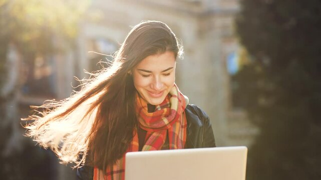 Young woman with long flowing hair and plaid scarf smiles while working on laptop outside in sunlight. Concept of technology and connection