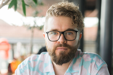 Close-up portrait of a man with glasses, curly hair, and beard, sitting outdoors at a cafe. Neutral...
