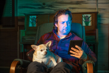 Obraz premium A man is sitting in a chair with a dog on his lap. He is holding a tablet in his hand reading a book.