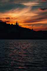 Contours city buildings and silhouette of a church at sunset with vibrant orange and purple hues over a calm river.
