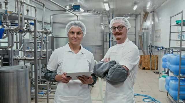 Medium arc portrait of two male and female Caucasian cheese making factory employees in white uniforms and caps standing in raw milk processing facility in front of machines, looking at camera