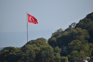 Turkish flag flying over a hilltop in Istanbul, surrounded by dense green trees and distant mountains under a clear blue sky
