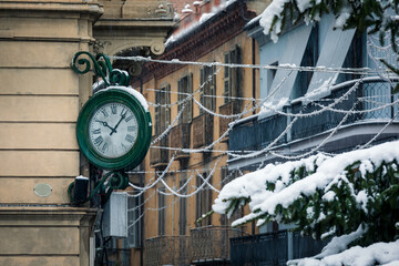 A vintage green clock with Roman numerals is mounted on the corner of a building in a snowy, italian town street.
