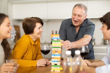 Smiling senior man or father having fun enjoying tower building game with wife and adult children at light comfortable family home