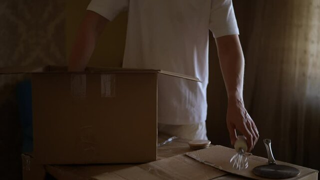 Closeup of man carefully placing pan into cardboard box, surrounded by other kitchen items and packing materials, during home relocation process. Concept of new life in new home relocation.