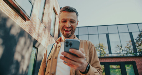 Bottom view of satisfied man loudly smiling after reading message on smartphone from friend or watching funny video. Enjoying communication online. Modern lifestyle. Spending time at street.