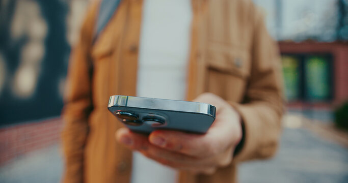 Camera view of man entering message with both hands on mobile phone. Actively typing text on touchscreen. Using digital gadget for remote communication. Standing at street in front of modern building. - Powered by Adobe