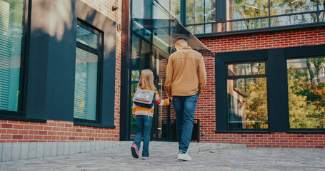 View from behind of Caucasian father and daughter walking together back home from school. Going towards entrance to modern building with big windows. Child carrying backpack. Sunny weather.